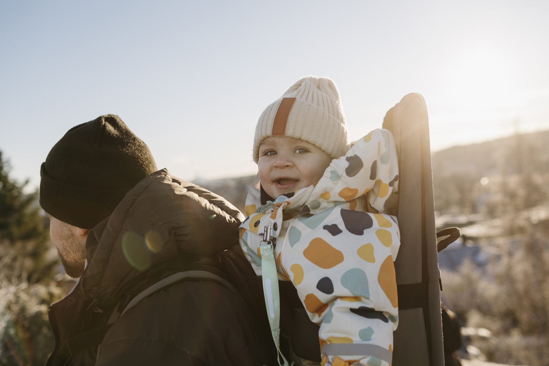Barn på under ett år sitter i en bæremeis og smiler mot kamera, faren ser vi bare bakhodet på